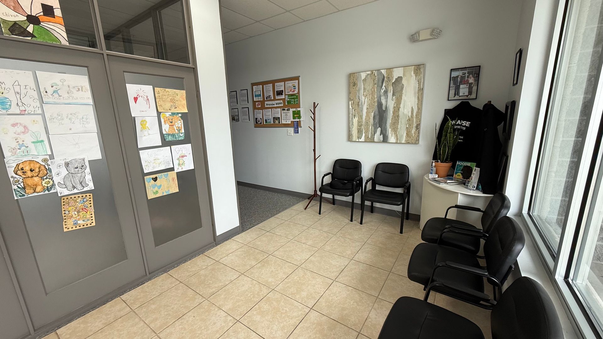 Waiting room with tiled floor, black chairs, and artwork. Grey doors on left, large window on right.