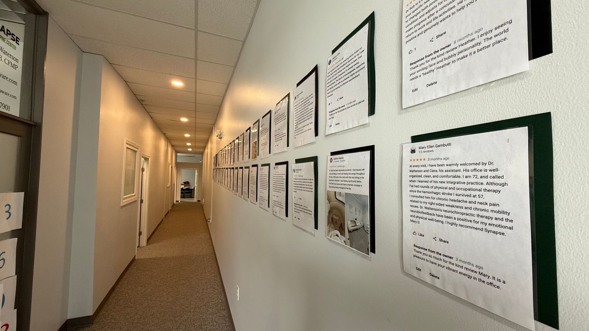 A narrow hallway with a white wall displaying framed documents. The floor is carpeted, and a door is at the end of the hallway.