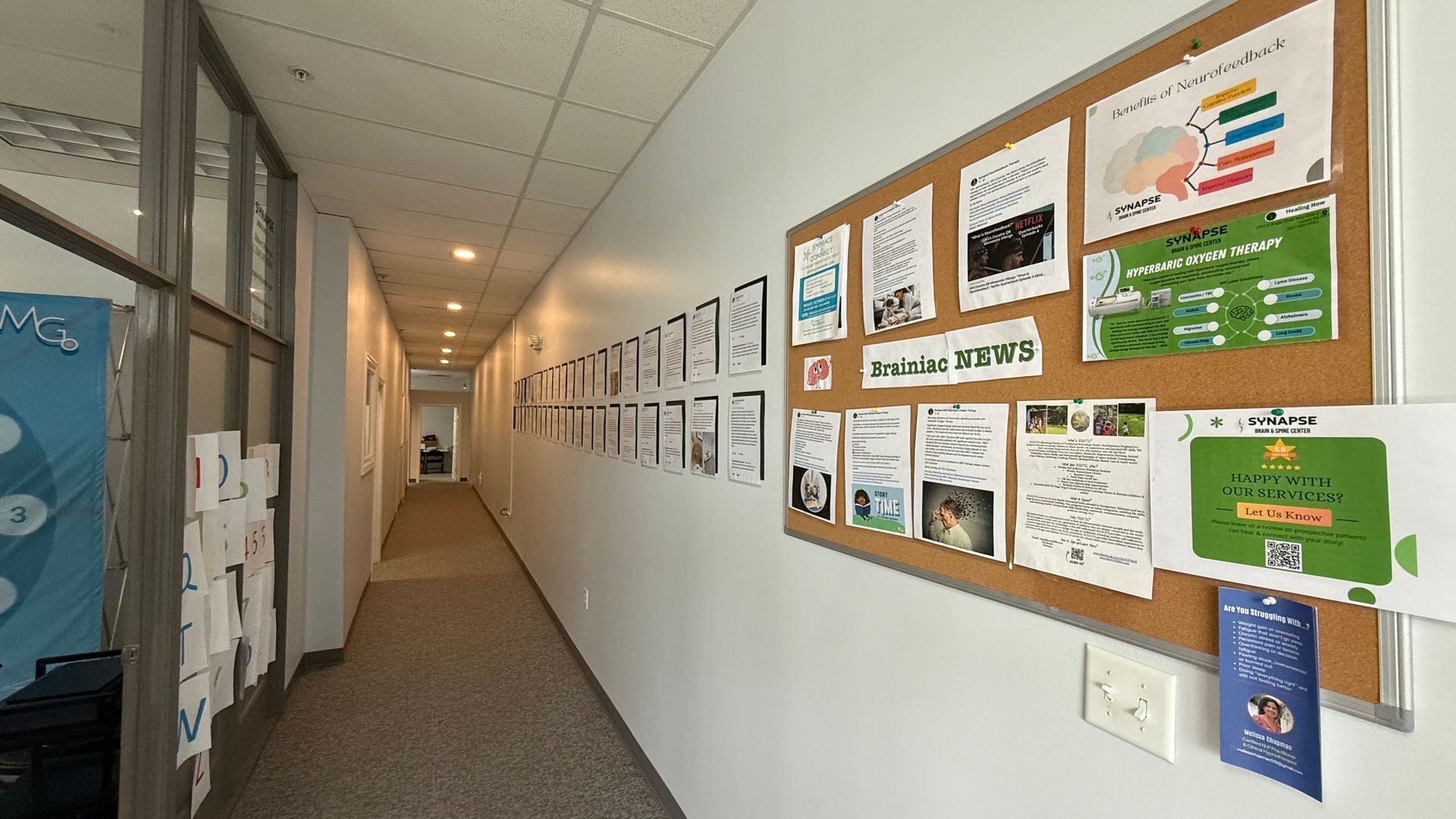 A long office hallway with white walls, a corkboard with papers, and framed art. The floor is carpeted.