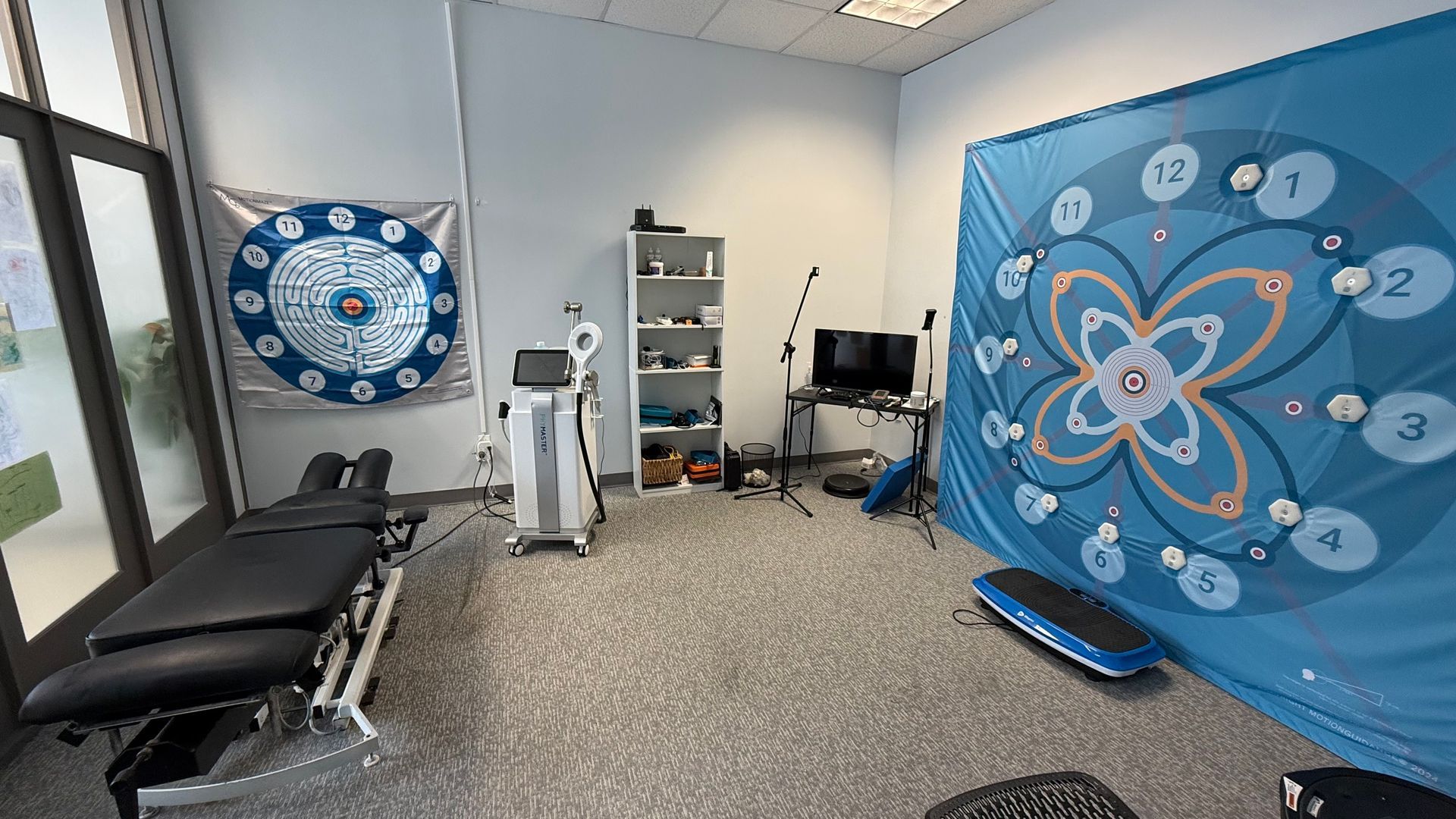 A therapy room with medical equipment, two blue patterned wall hangings, and a massage table.