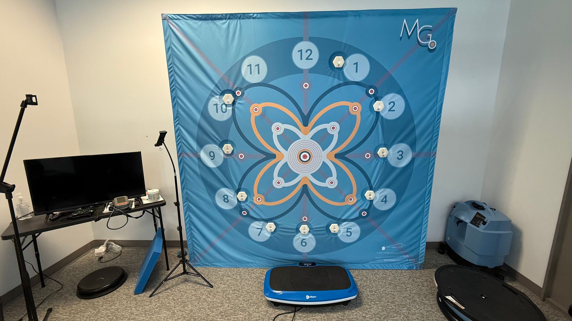 A room features a large blue wall graphic with a circular design, numbers, and a floral pattern. Several exercise machines are in front of it, with a desk and equipment to the side.