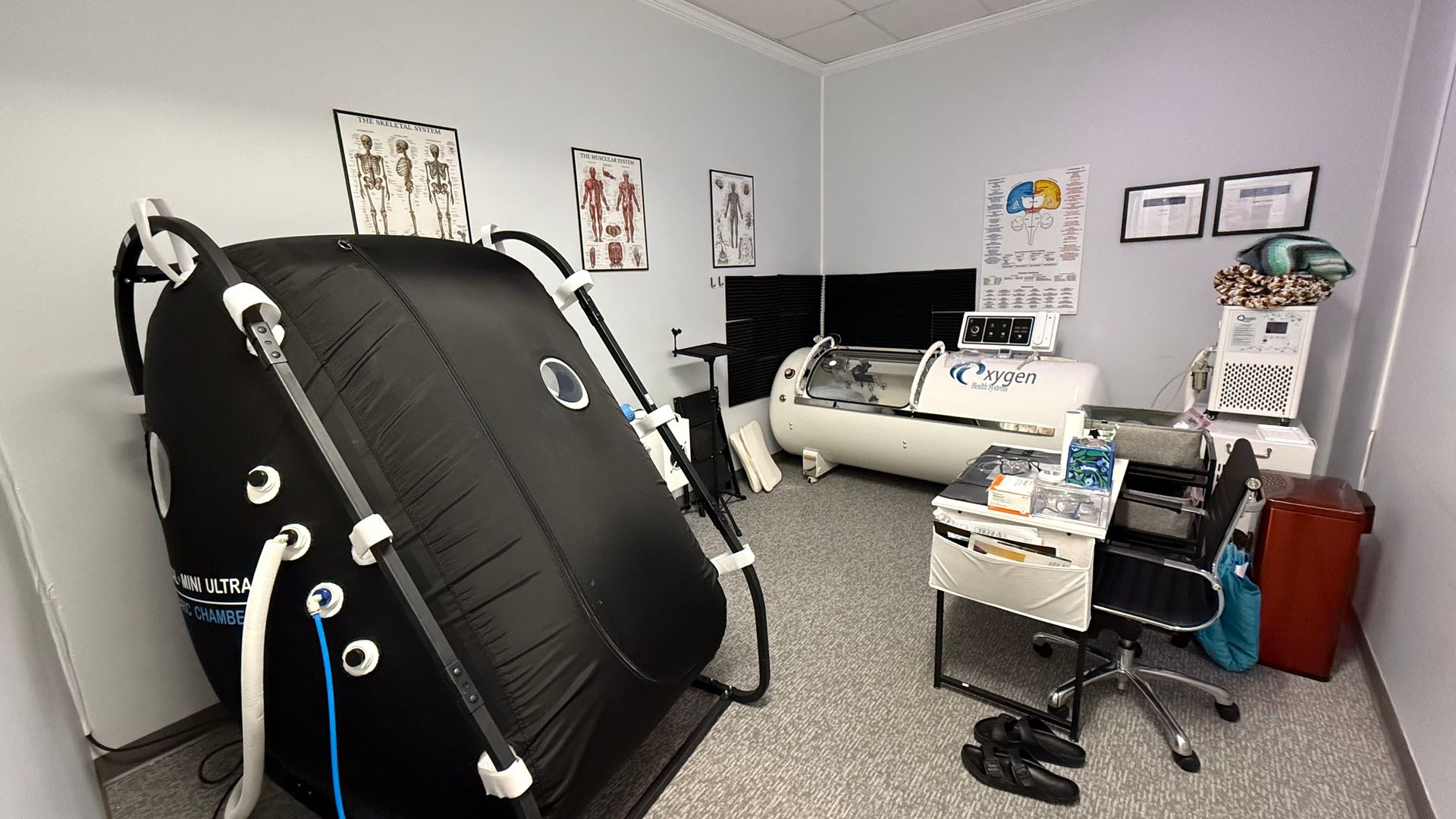 A medical room with a black, inflatable hyperbaric chamber and a white, hard-shelled chamber. Skeletal charts hang on the wall.