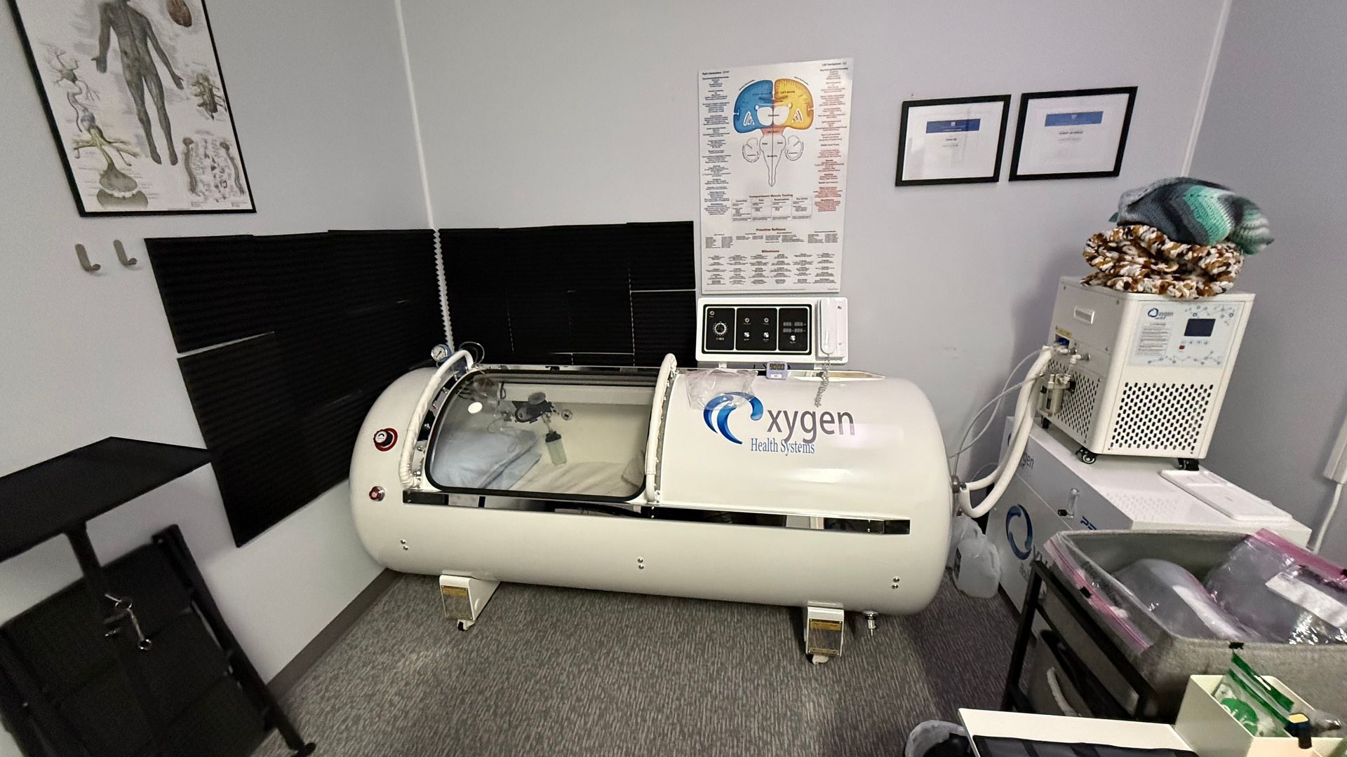A white hyperbaric oxygen chamber in a medical room. The chamber is open, revealing a bed. There's a control panel and supporting equipment nearby.