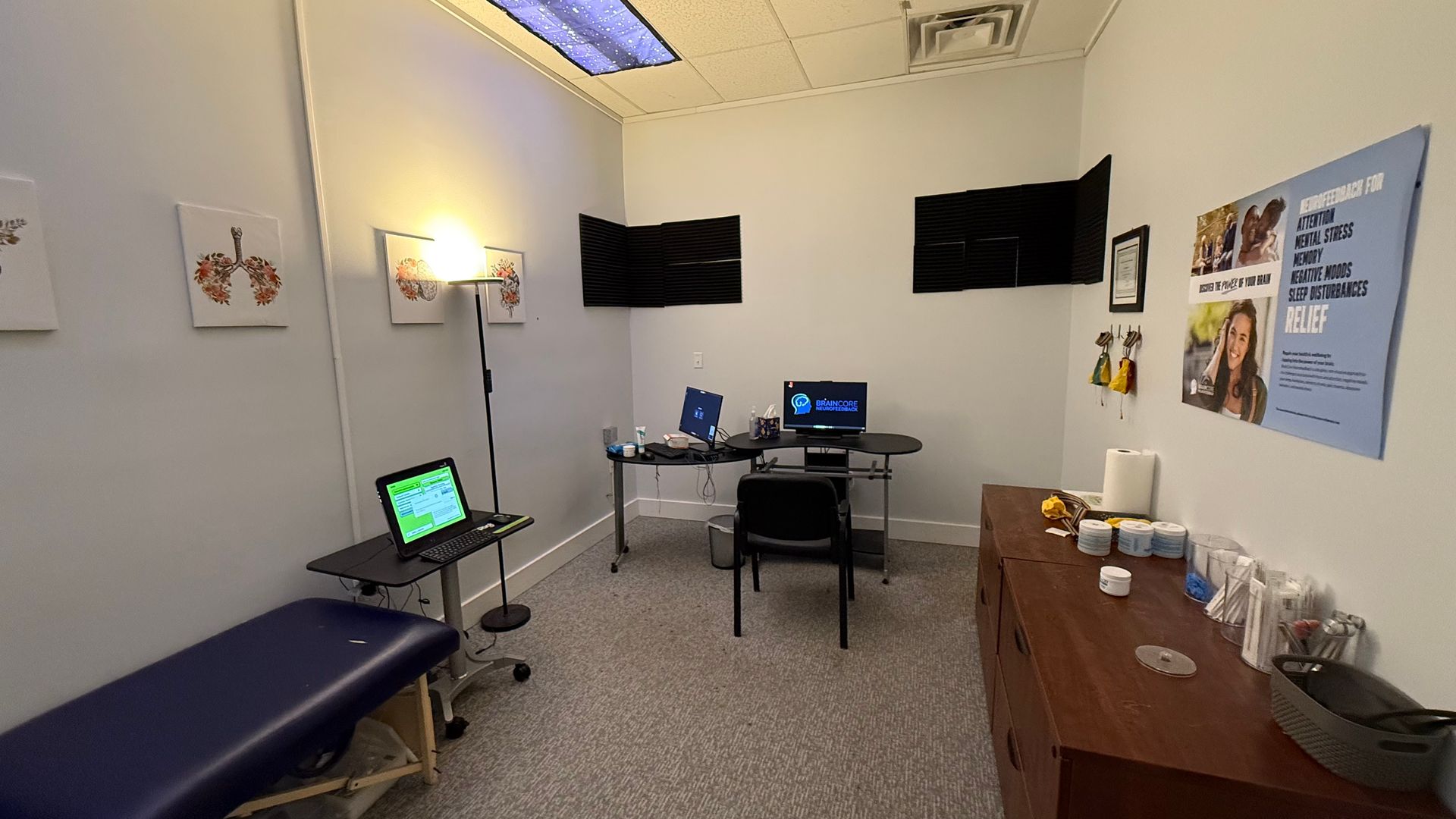 Medical exam room: blue exam table, two monitors on the wall, computer, and desk. Walls are white, carpet is gray and brown.