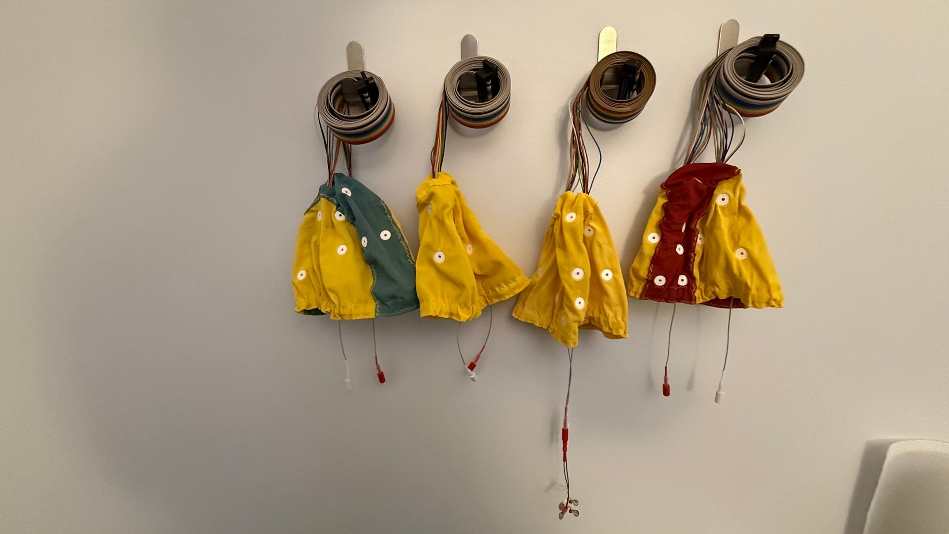 Four colorful fabric pouches hang from metal fixtures on a white wall, with red paint drips below.