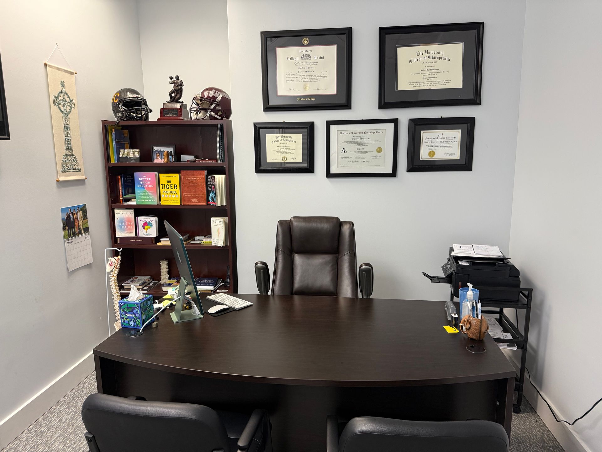 A dark brown office desk with a leather chair faces a bookshelf and framed certificates on a light gray wall.