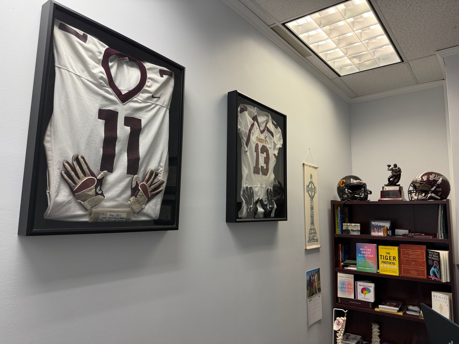Wall-mounted football jersey and photograph, alongside a shelf with helmets and books, in an office.