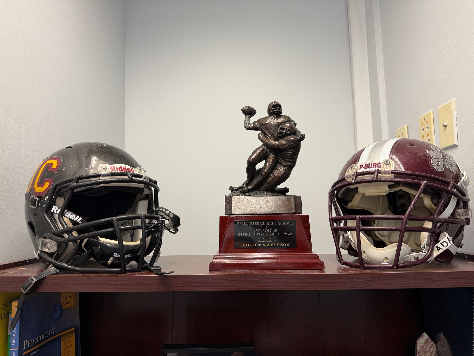 Two football helmets flanking a trophy on a shelf. A dark helmet with a