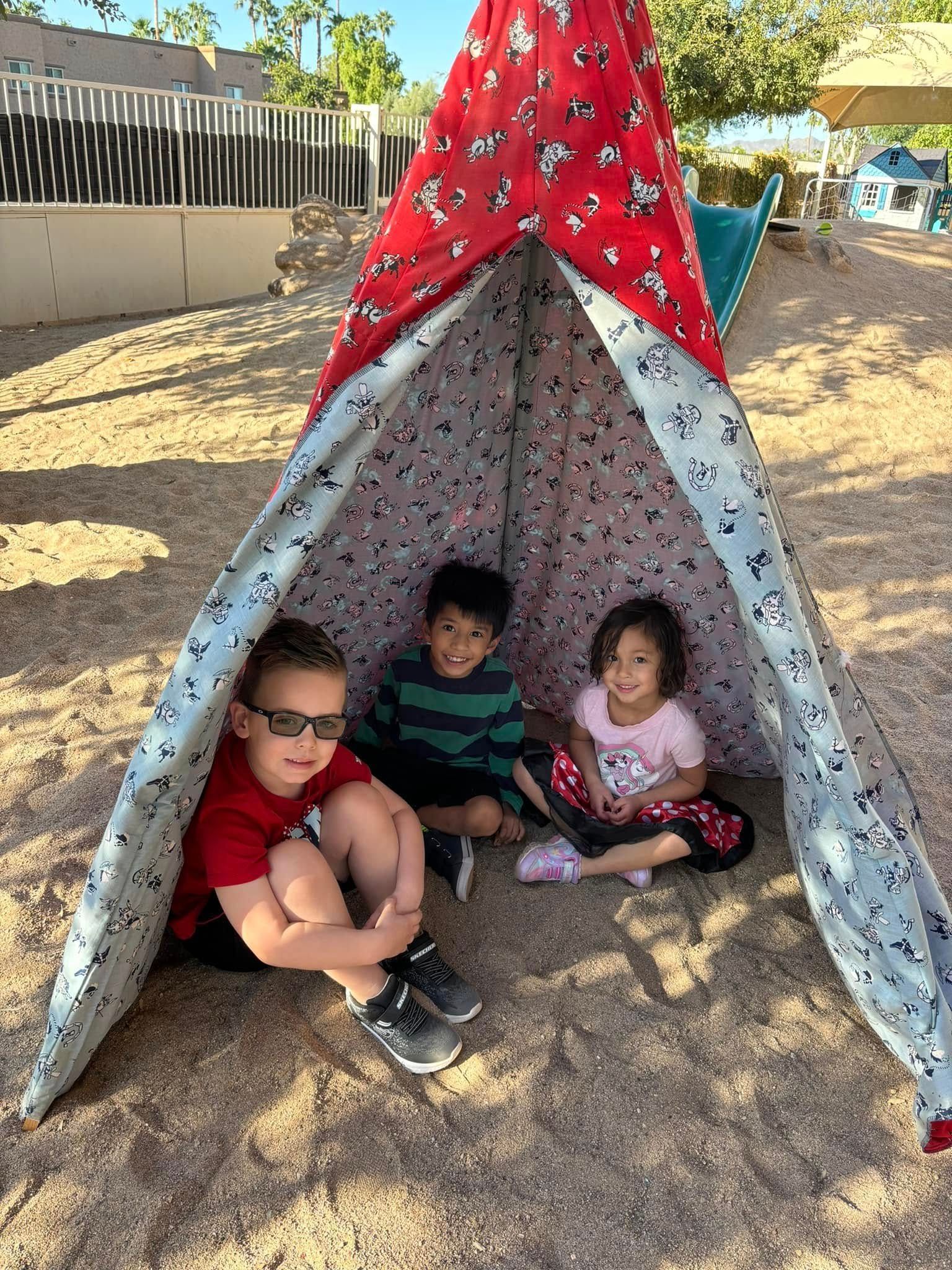 Three children sit inside a red, white and blue patterned play tent on a playground.