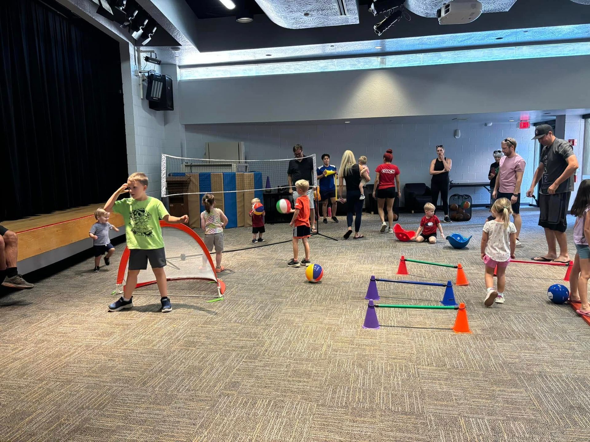 Children playing games in a large room with adults. Soccer nets, cones, and obstacles are visible.