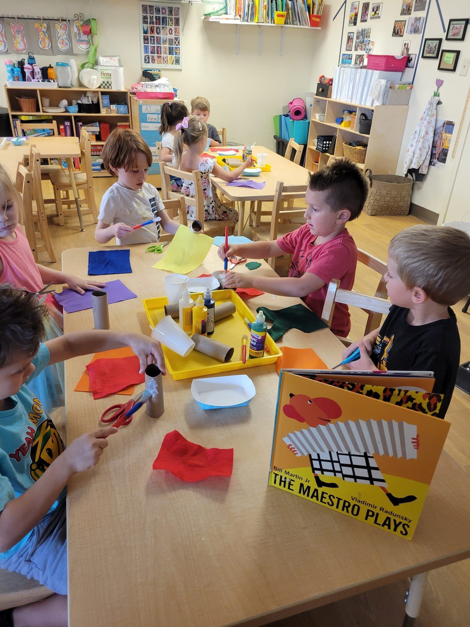 Children at a table, crafting with paper, paint, and cardboard tubes in a colorful classroom setting.