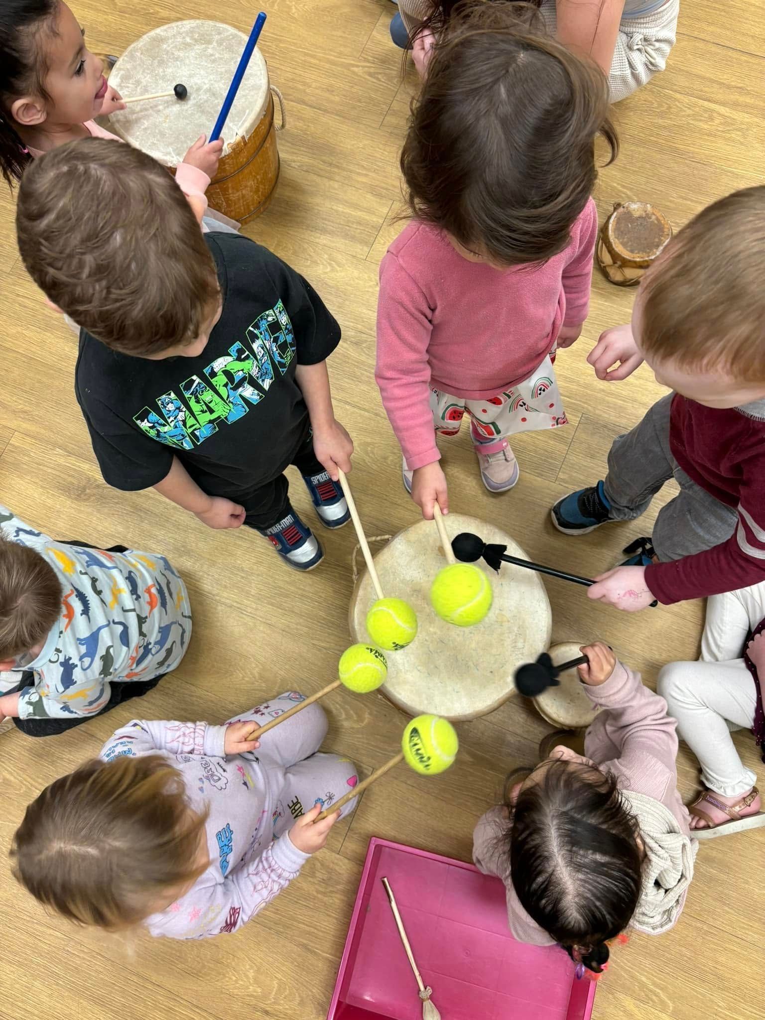 Children playing drums in a circle, using drumsticks. Yellow balls rest on the drums.
