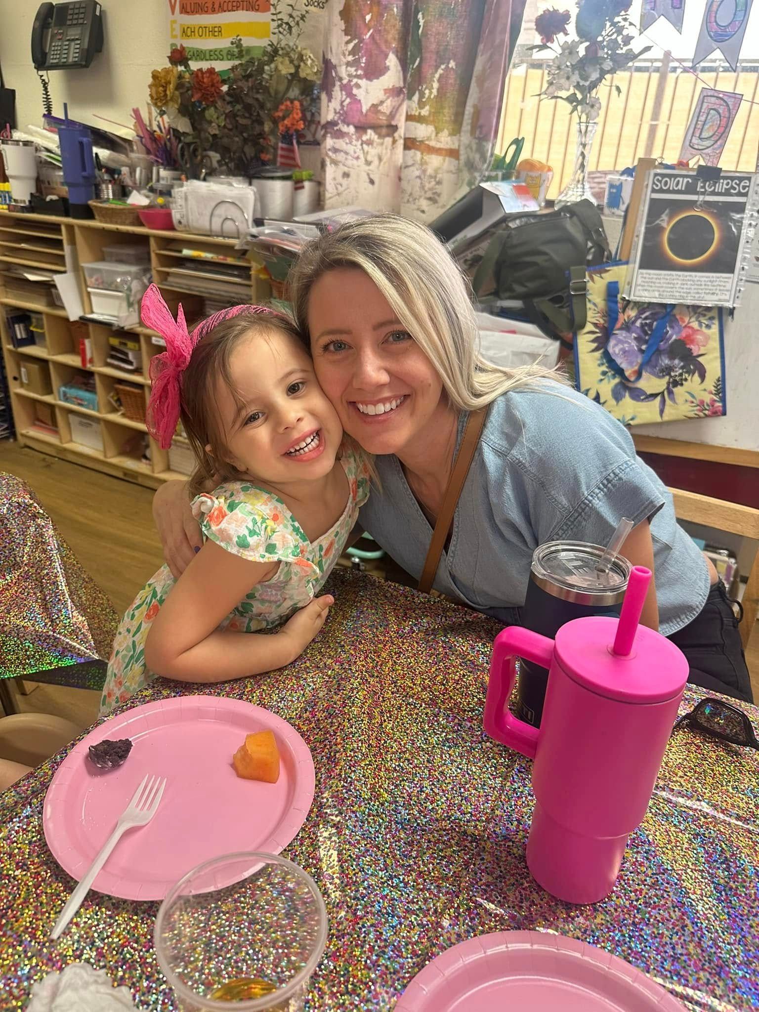 Woman and child smiling, hugging at a colorful table; drink and plates nearby.