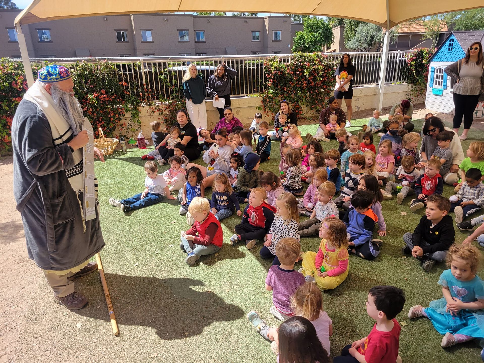Group of children seated on grass, listening to a person in traditional clothing, outdoors under a canopy.