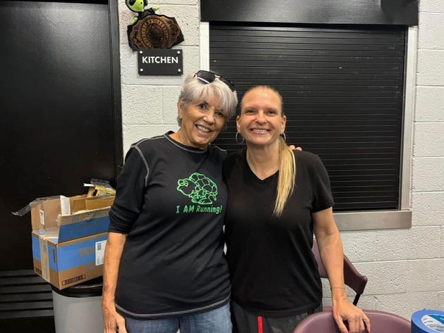 Two women smiling, arm in arm, in a kitchen. One wears a black shirt with a green turtle, the other has a ponytail.