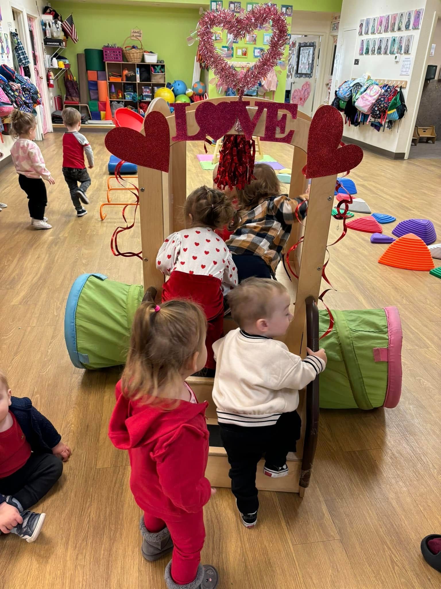 Children climb a wooden structure decorated for Valentine's Day. Red and pink hearts are featured.