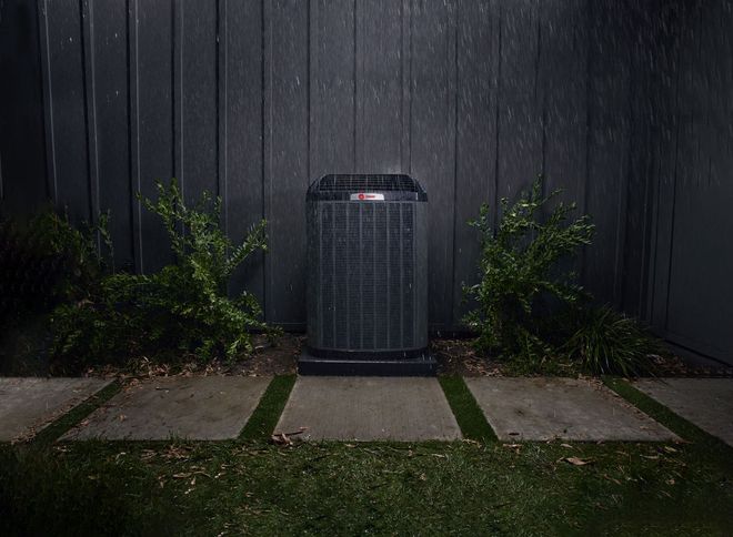 Outdoor air conditioner unit against a dark fence, flanked by bushes and stepping stones.
