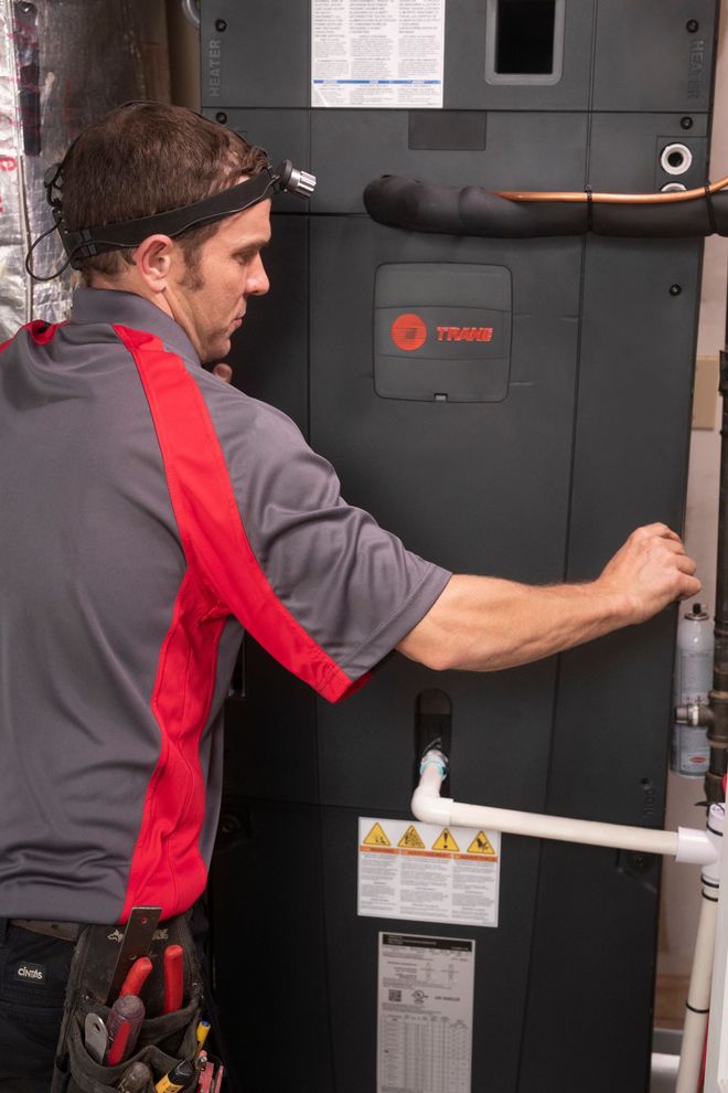 HVAC technician in a yellow hard hat repairs an air conditioner unit outside a building.