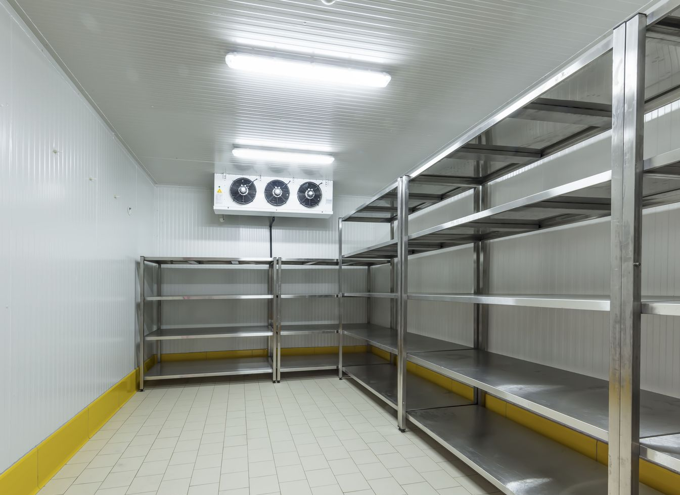 Empty commercial walk-in refrigerator with metal shelves, white walls, ceiling lights, and ventilation unit.