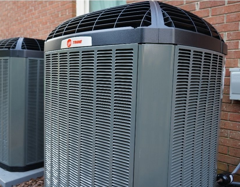 Two Trane air conditioning units, gray with black tops, against a brick wall.