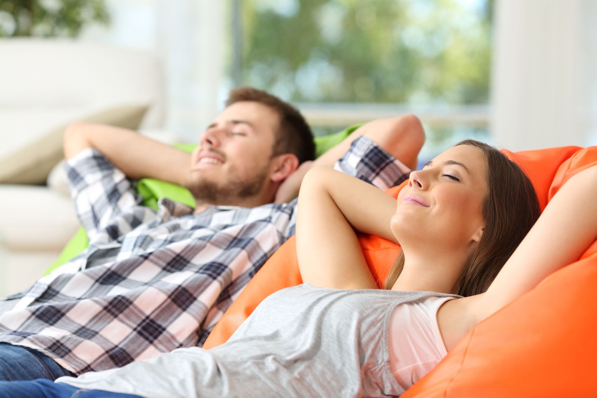 A couple relaxing on colorful beanbag chairs, hands behind heads, eyes closed, enjoying a sunny living room.