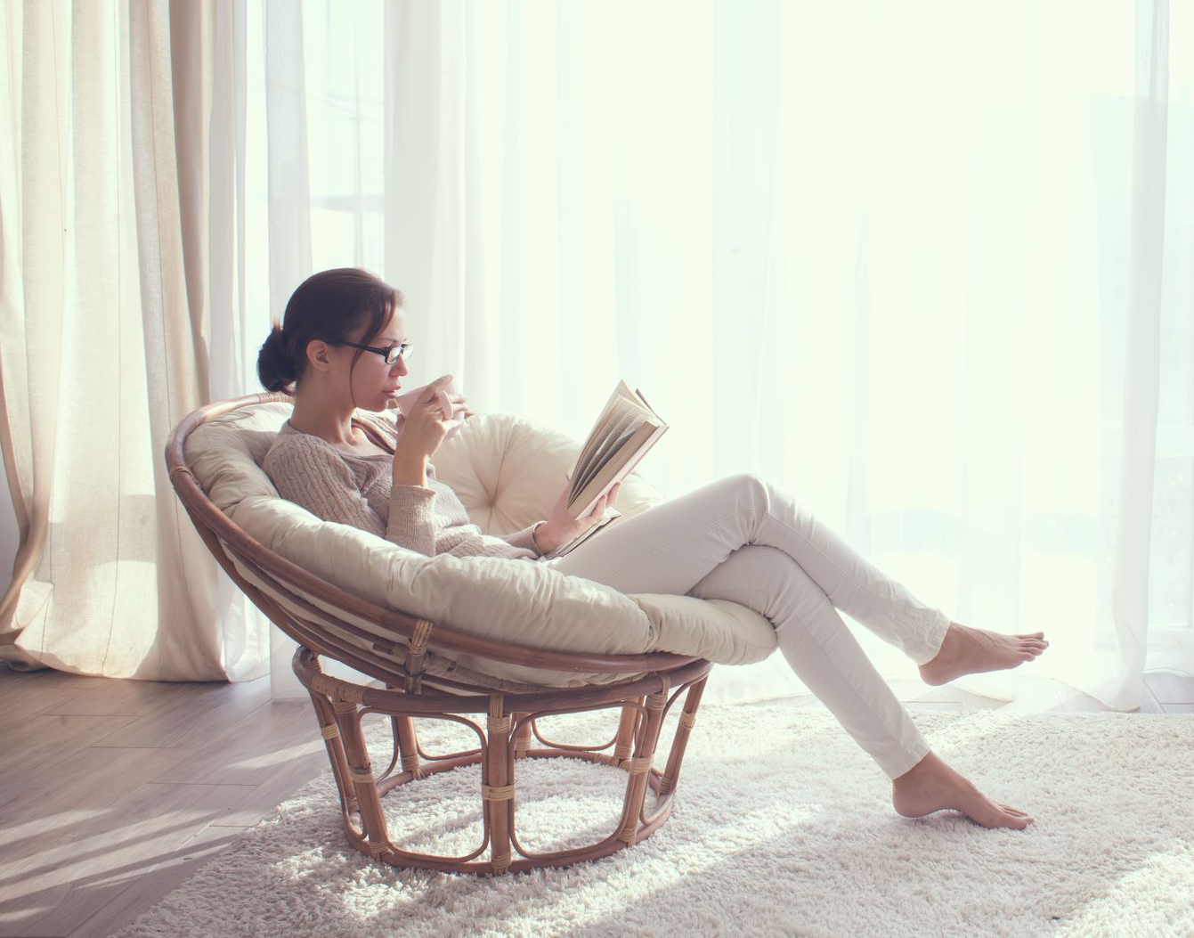 Woman reading, sitting in a round chair near a bright window with white curtains, sipping from a cup.