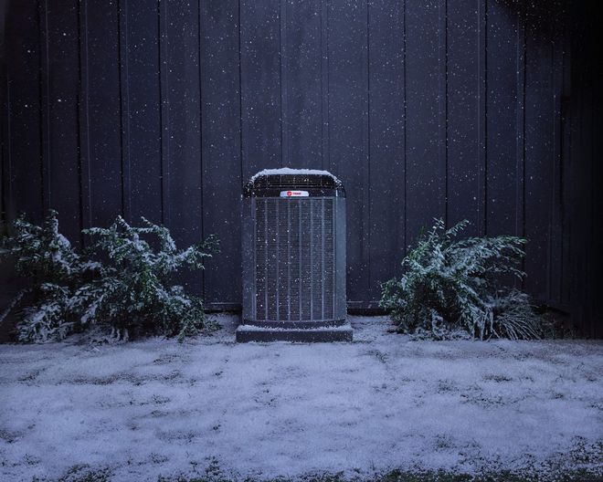 Snowy yard at night with a centered outdoor air conditioner unit against a dark wall.