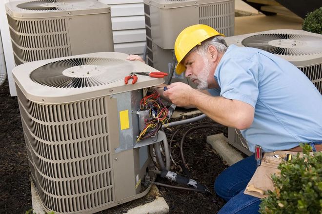 HVAC technician in a yellow hard hat repairs an air conditioner unit outside a building.