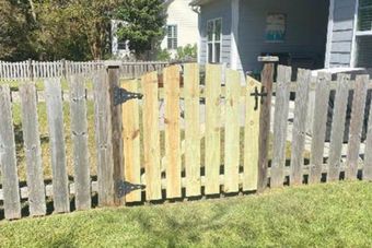 A wooden gate in a picket fence with new, light-colored wood set against weathered gray