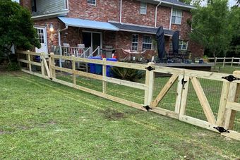 A wooden fence with two gates surrounds a grassy yard with a brick house in the background