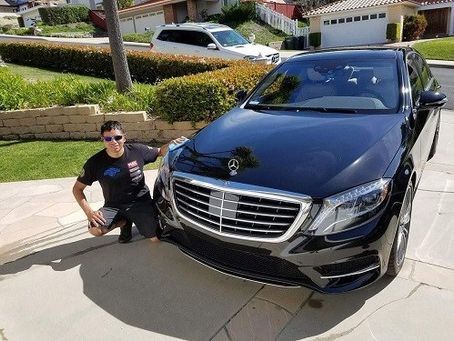 A person in a black t-shirt squats beside a shiny black Mercedes-Benz sedan parked in a sunlit residential driveway.