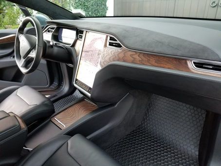 The interior of a Tesla with dark leather seats, wood trim, a large central touchscreen, and honeycomb floor mats.