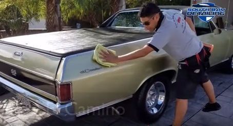A person in a gray shirt wipes the back panel of a vintage gold El Camino SS vehicle parked on a paved driveway.
