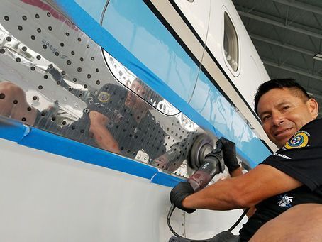 A technician in a uniform uses a power sander on the side of a white aircraft marked with blue tape.