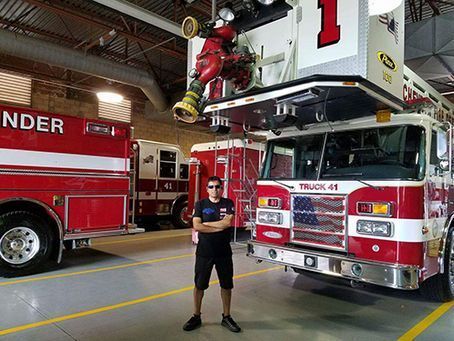 A person stands with arms crossed in a fire station between two red fire trucks, one labeled