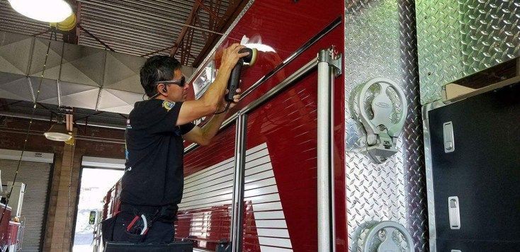 A person wearing sunglasses and a uniform uses a tool to work on the side of a red fire truck in a station.