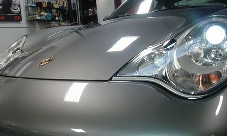 A close-up of a silver Porsche’s hood and headlight in a garage setting with bright, reflective lighting.