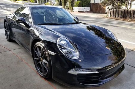 A sleek, black Porsche 911 parked on a paved driveway near a street and wooden fence.
