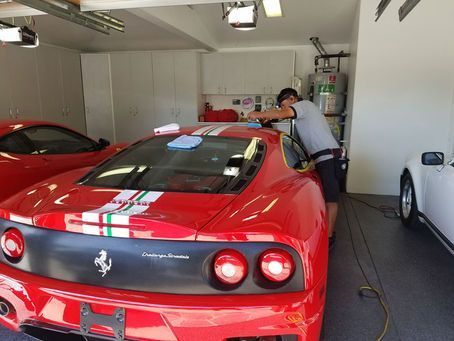 A person in a gray shirt polishes a red sports car with a racing stripe in a garage with other cars.