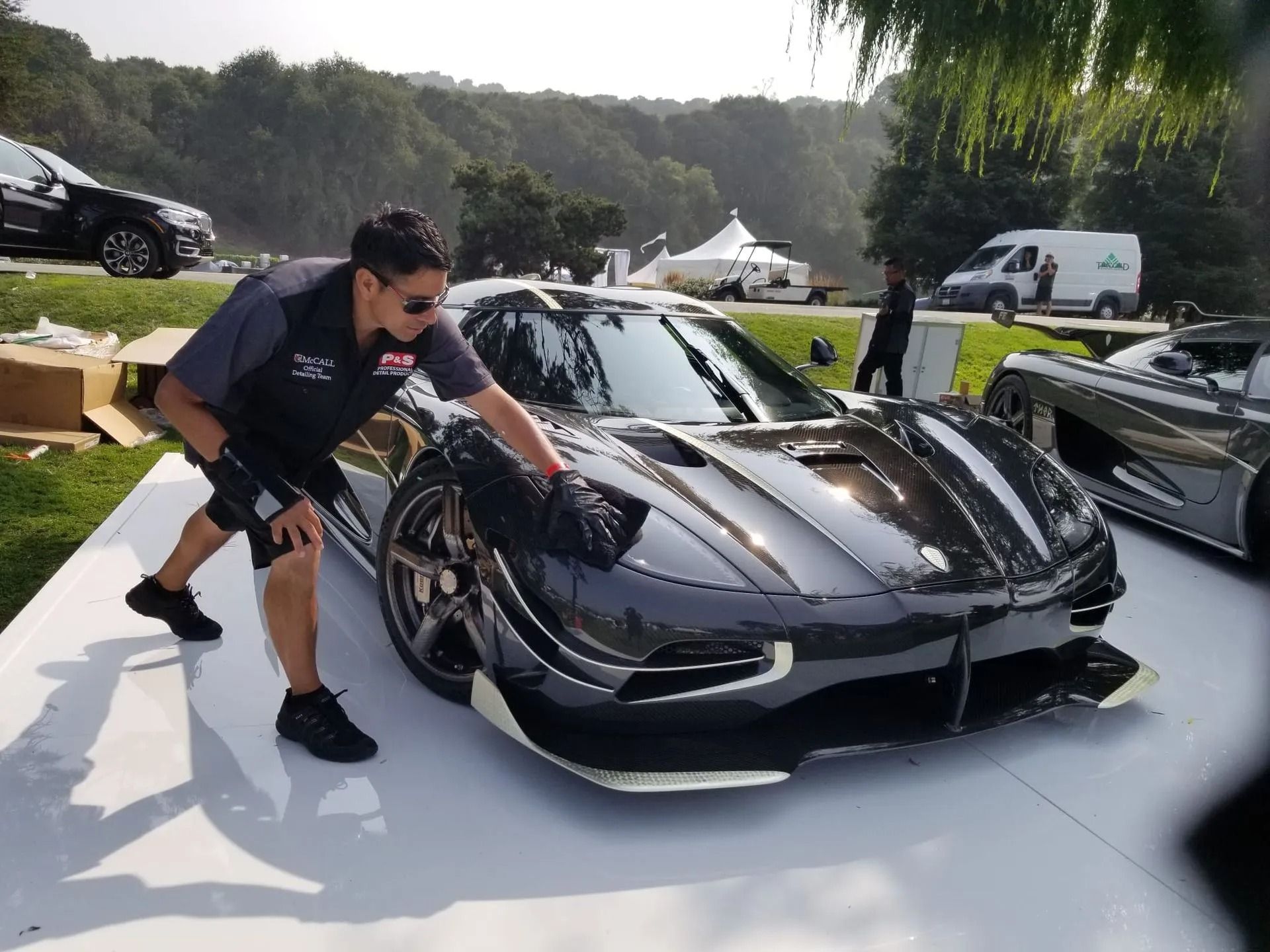 A worker polishes a dark, sleek sports car on a white platform at an outdoor event.
