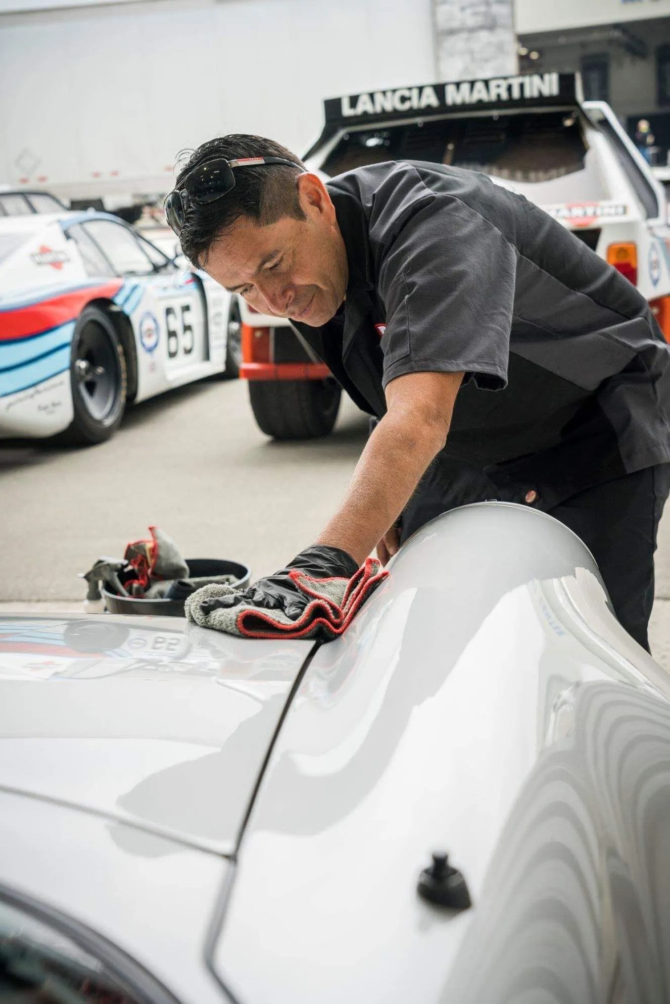 A person in a dark uniform wipes down a white race car at a track with Lancia Martini vehicles in the background.