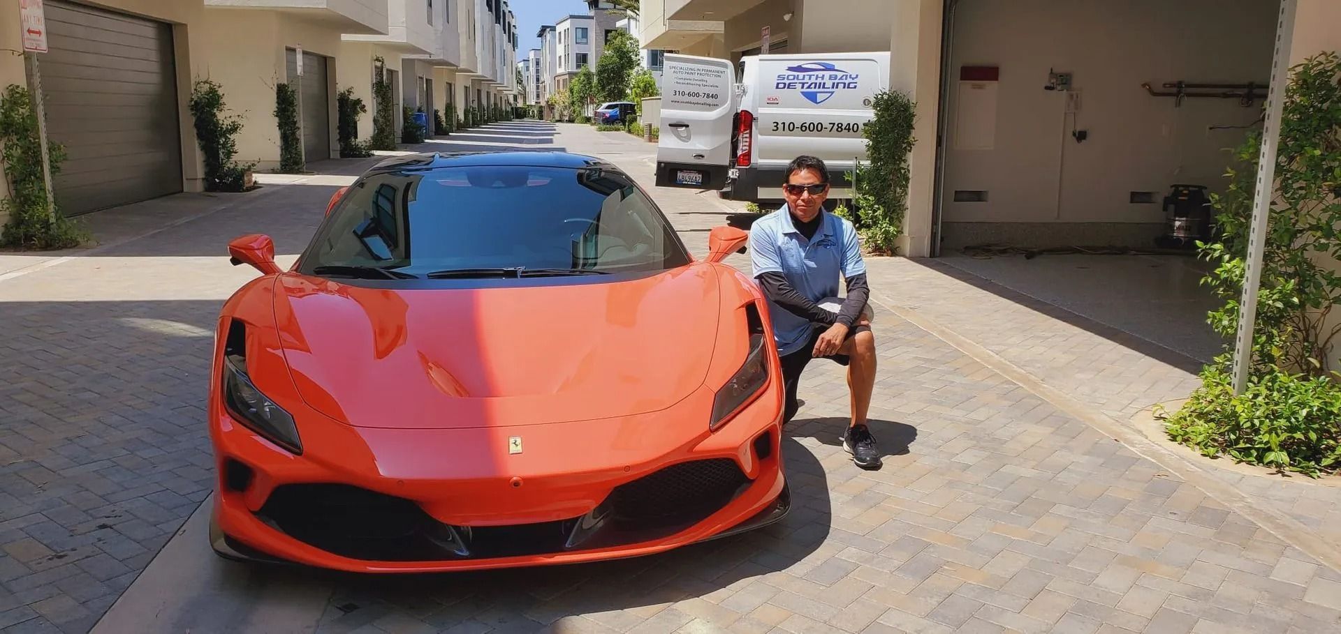 A person kneels next to a bright orange sports car parked on a paved driveway between two buildings.