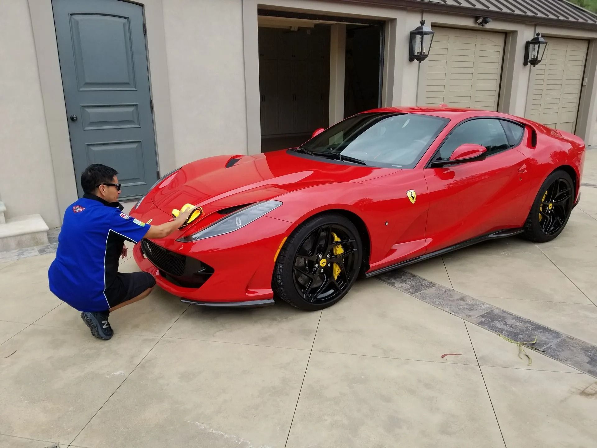 A person in a blue shirt wipes the hood of a red Ferrari sports car parked on a concrete driveway near a garage.