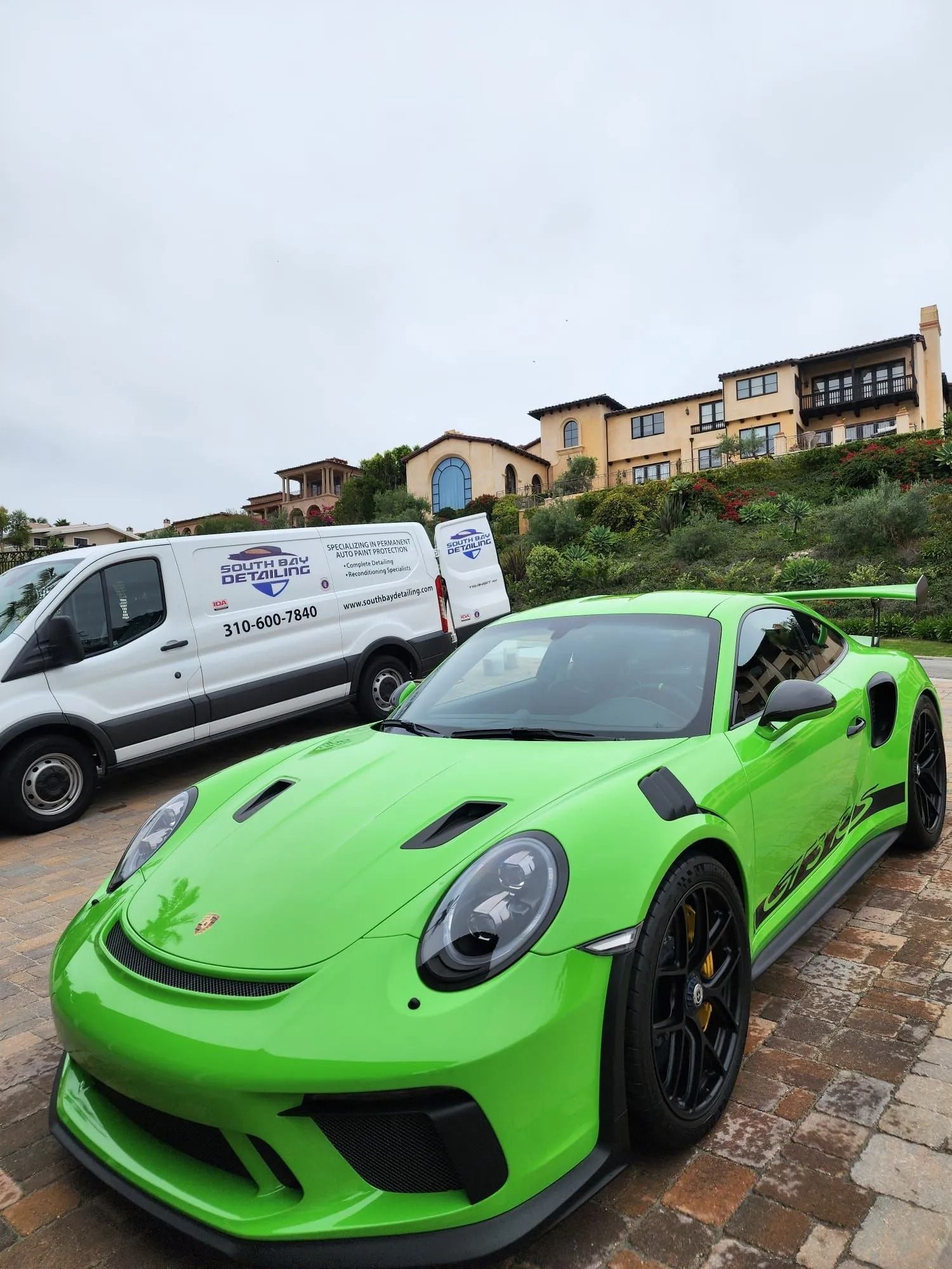 A bright green Porsche 911 GT3 RS parked on a paved driveway in front of a white utility van and a large suburban home.