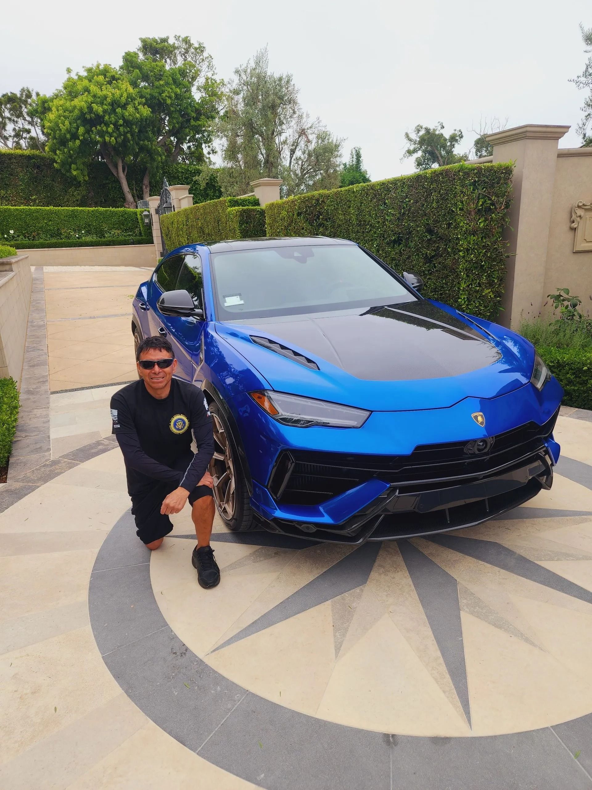 A person in dark clothing kneels beside a vibrant blue Lamborghini Urus SUV on a circular stone driveway.