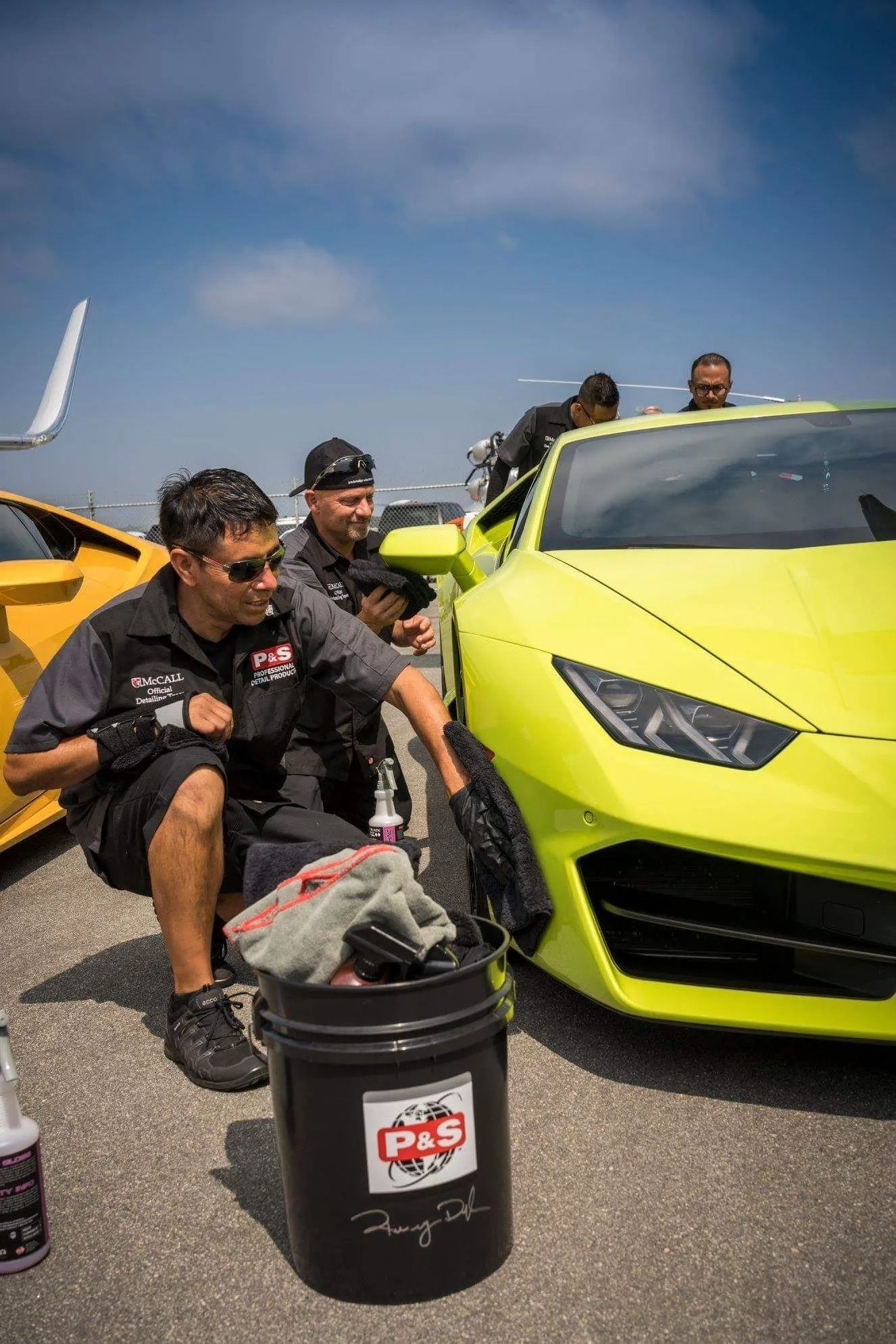 A person kneels while detailing a bright lime-green sports car parked outdoors near a bucket with a P&S brand label.