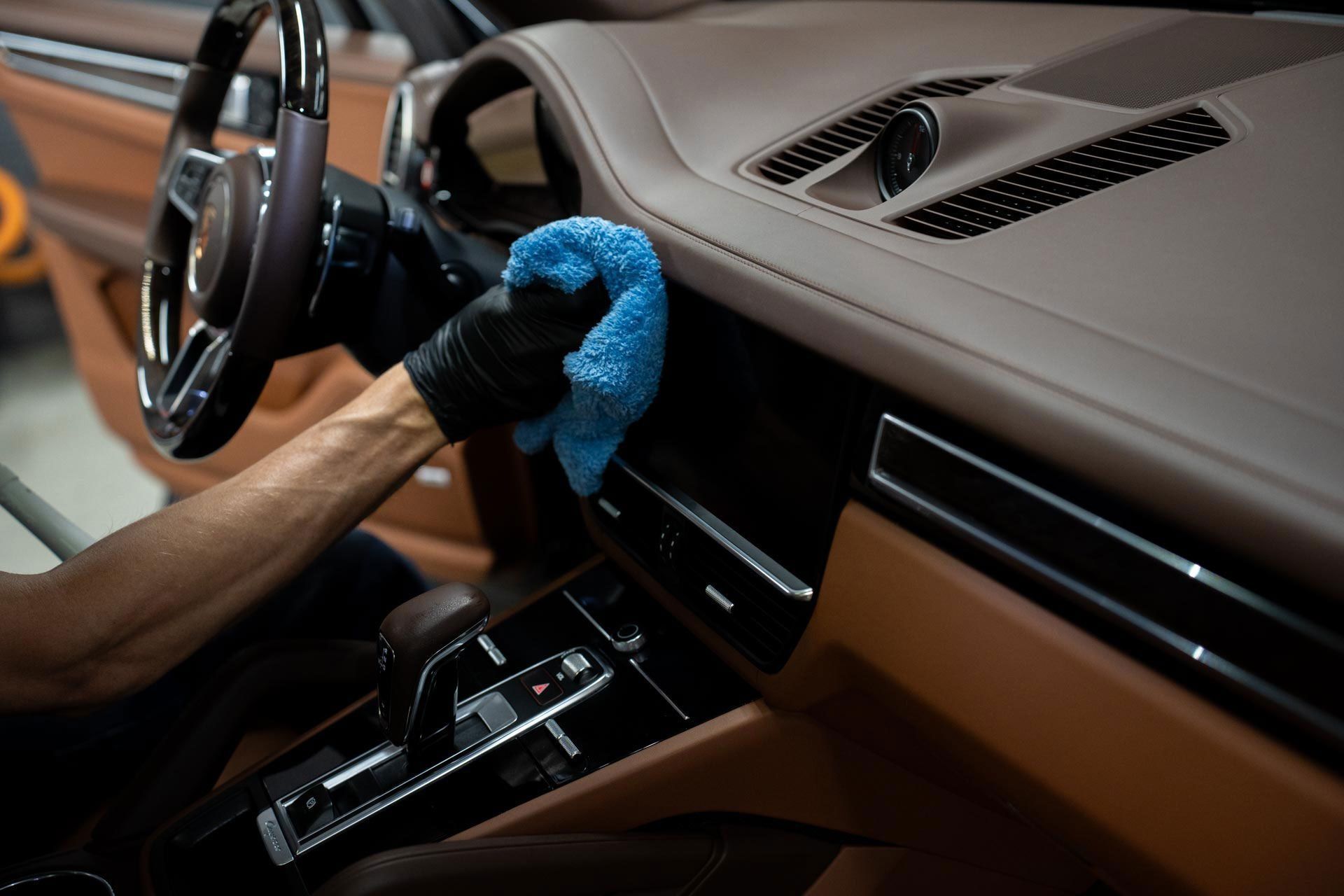 A person wearing black gloves wipes a vehicle's dashboard with a blue microfiber cloth in a tan-and-black car interior.