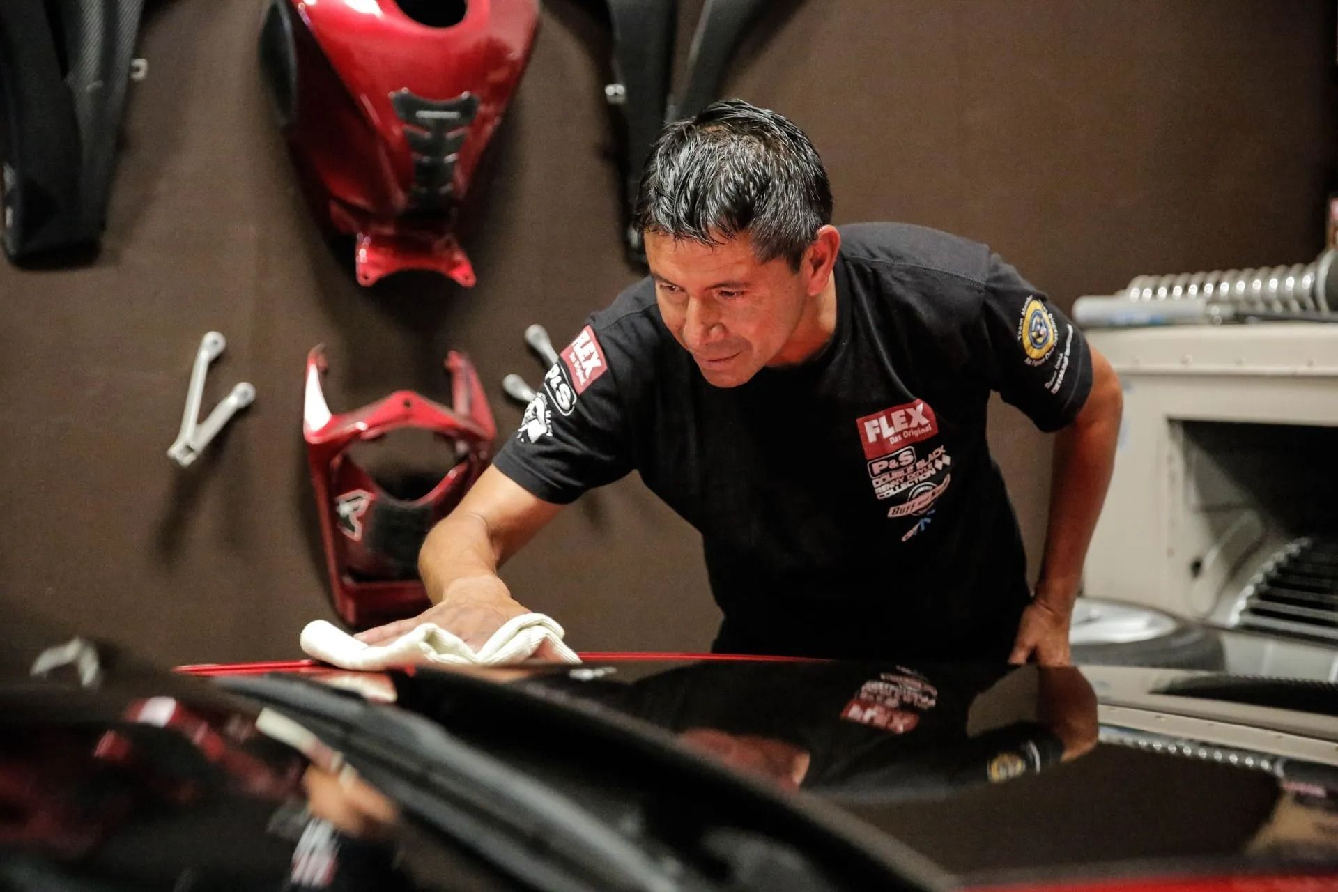 A mechanic in a black uniform wipes the hood of a red vehicle in a workshop with motorcycle parts on the walls.