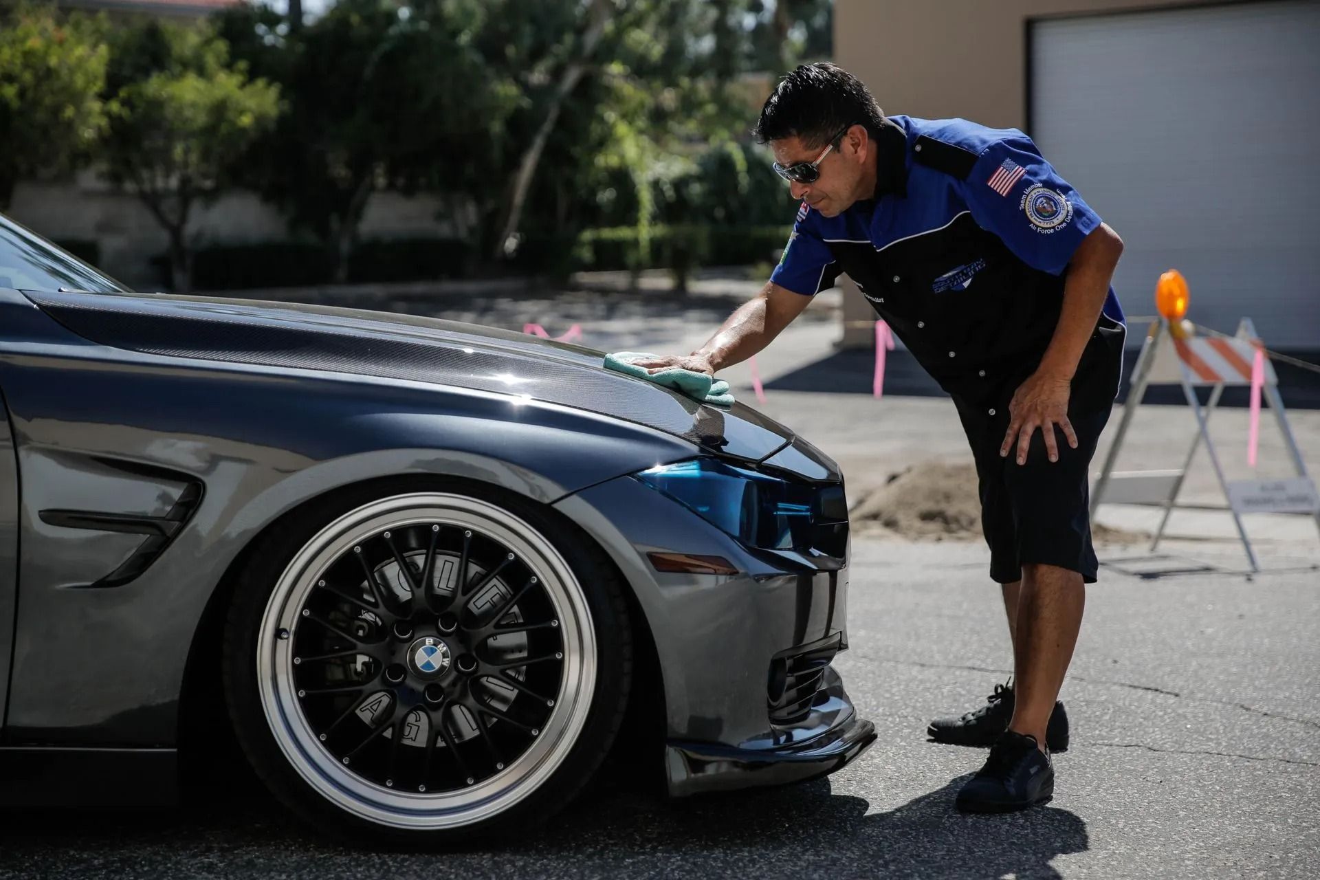 A person in a blue and black uniform polishes the hood of a dark grey BMW car in an outdoor setting.