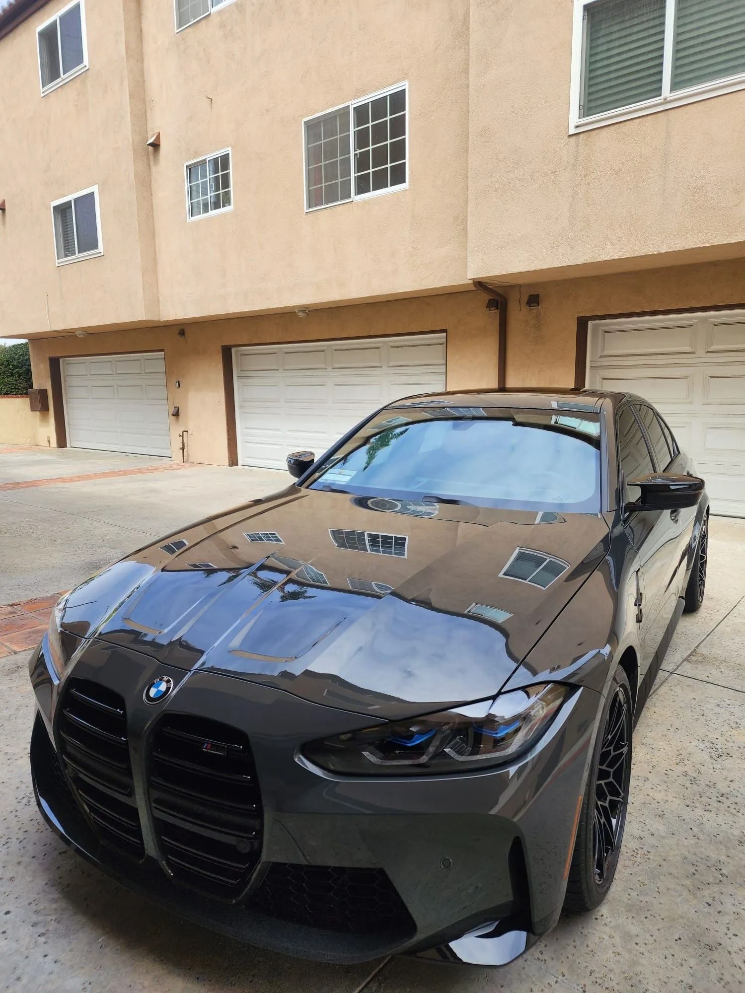 A dark grey BMW M3 sedan parked in a driveway in front of a tan residential building.